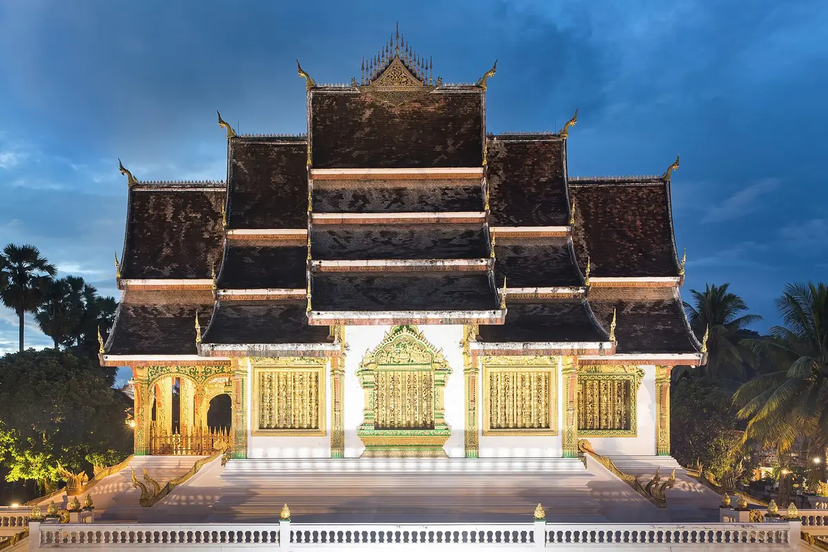 The tiered roofs and white terrace of Haw Pha Bang in Luang Prabang at blue hour, with the temple lit gold along its base.