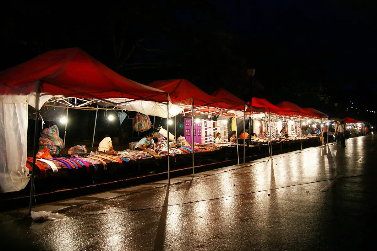 A row of red-roofed stalls selling textiles at the Luang Prabang night market, lit by bulbs on a wet street.