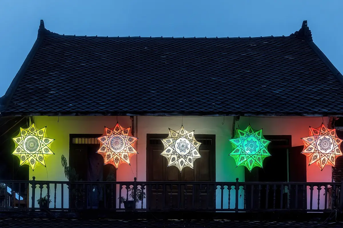 Five star-shaped paper lanterns glowing yellow, orange, white and green on the balcony of a wooden house at blue hour.