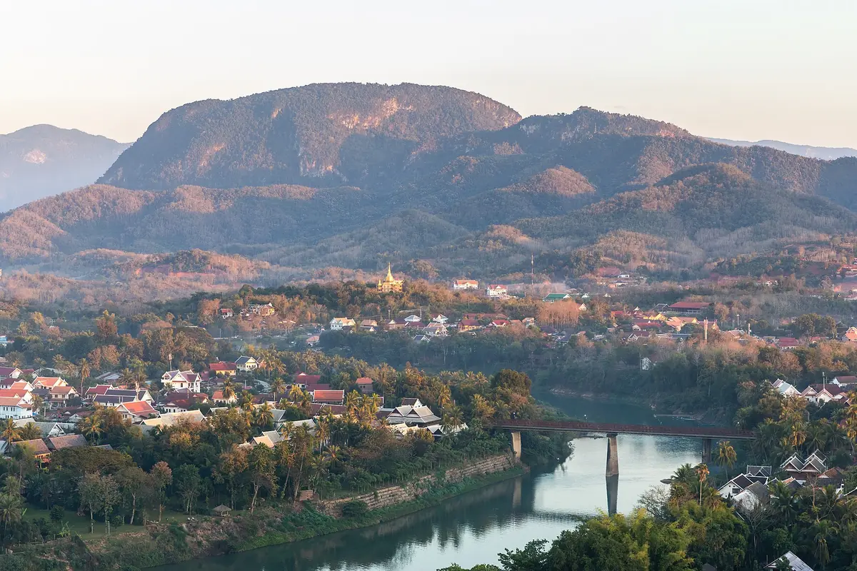Mountains, red-roofed houses and a bridge over the Nam Khan river in Luang Prabang, seen from Mount Phou Si at sunset.