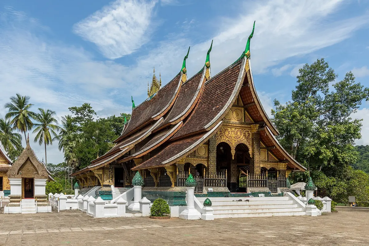 The multi-tiered, sweeping roofs of the sim at Wat Xieng Thong, with a gilded front gable, palms and a smaller shrine to the left.