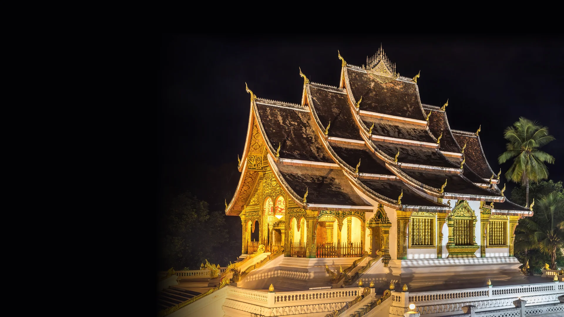 The gilded, three-tiered roofs of Haw Pha Bang in Luang Prabang, lit against the night sky with palm trees on the right.